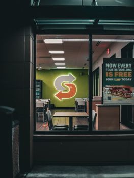 A dimly lit restaurant interior with a glowing neon logo sign, viewed from outside at night.