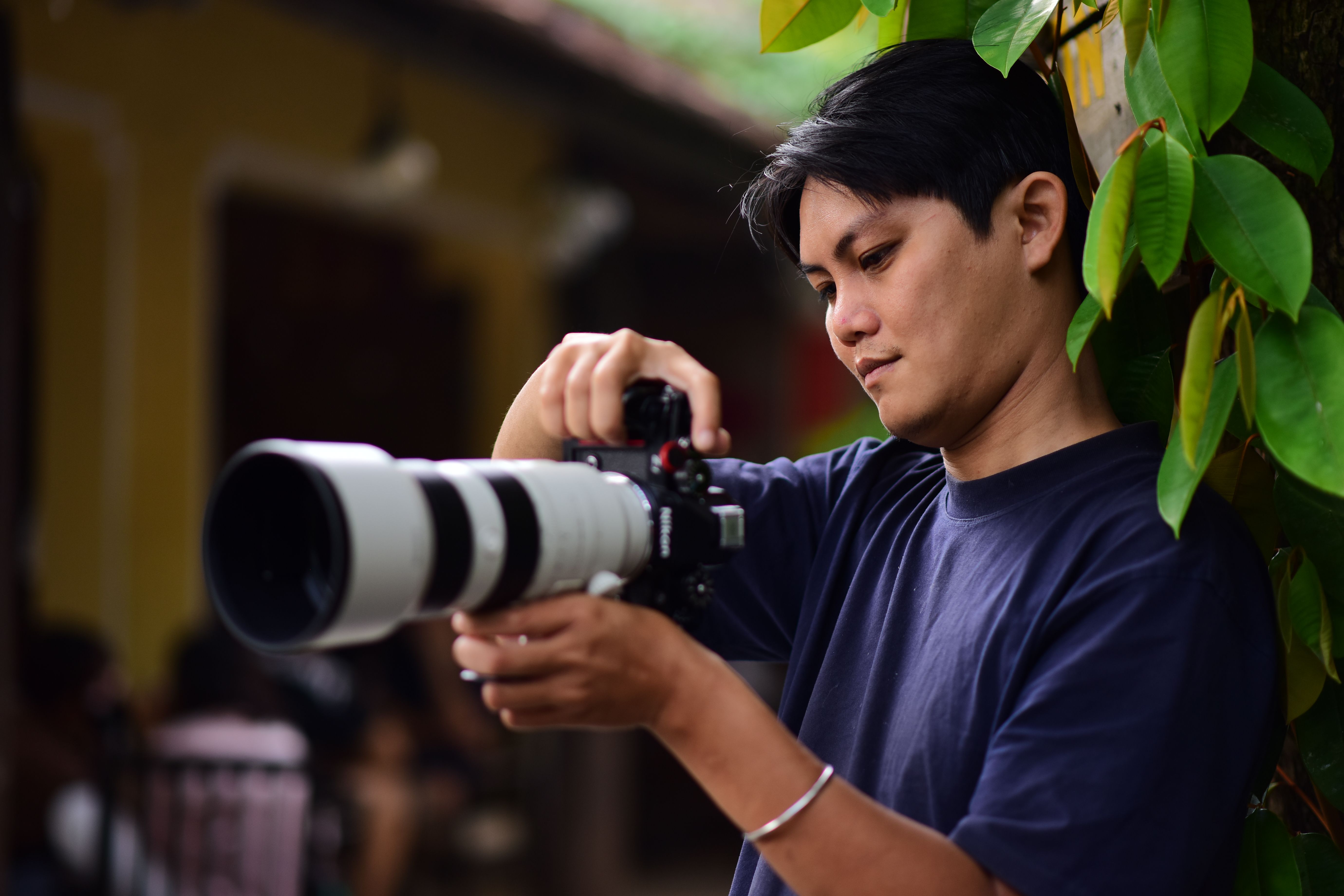 A focused photographer adjusts a professional camera outdoors, surrounded by greenery.
