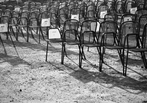 Rows of empty metal chairs casting shadows on a gravel surface in monochrome.