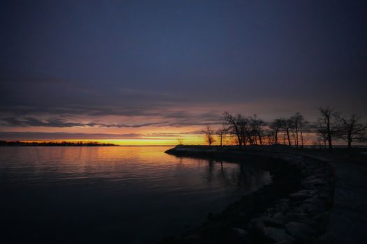 Peaceful lakeside sunset with silhouetted trees and vibrant sky reflection.