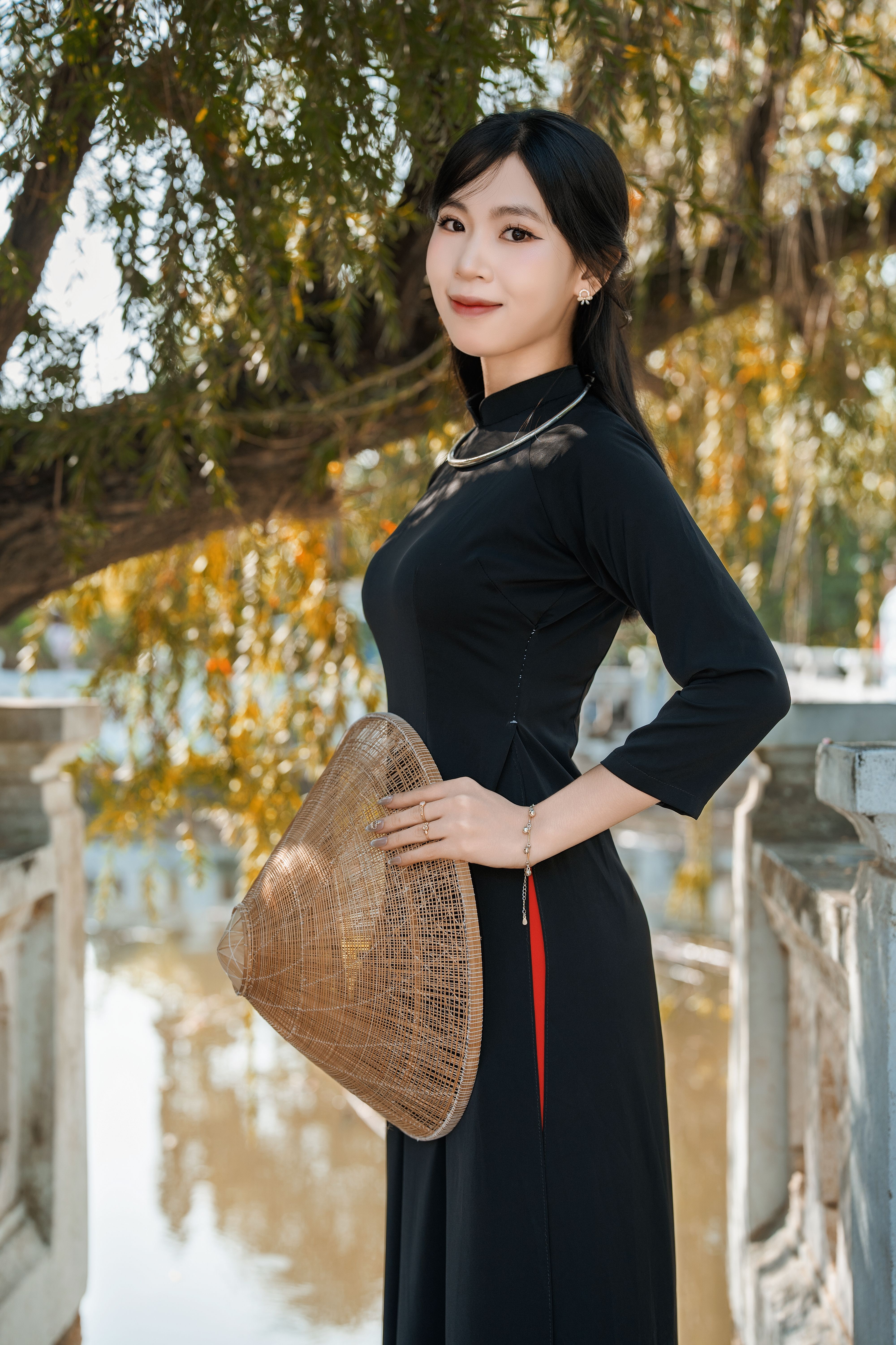 Woman in traditional Vietnamese dress holding a conical hat outdoors by a calm water body.