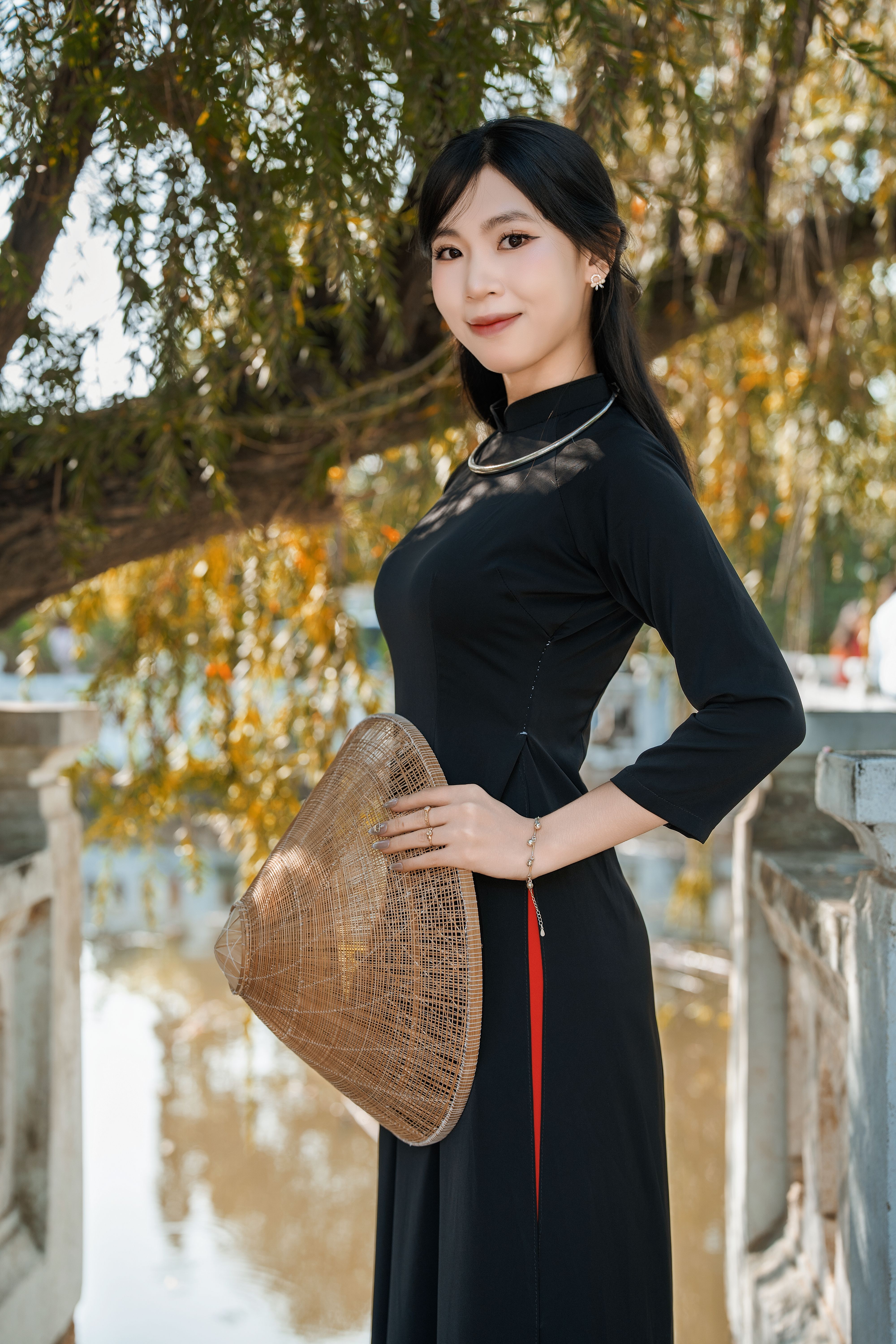 Young woman in black dress holding a traditional hat by a pond in autumn.