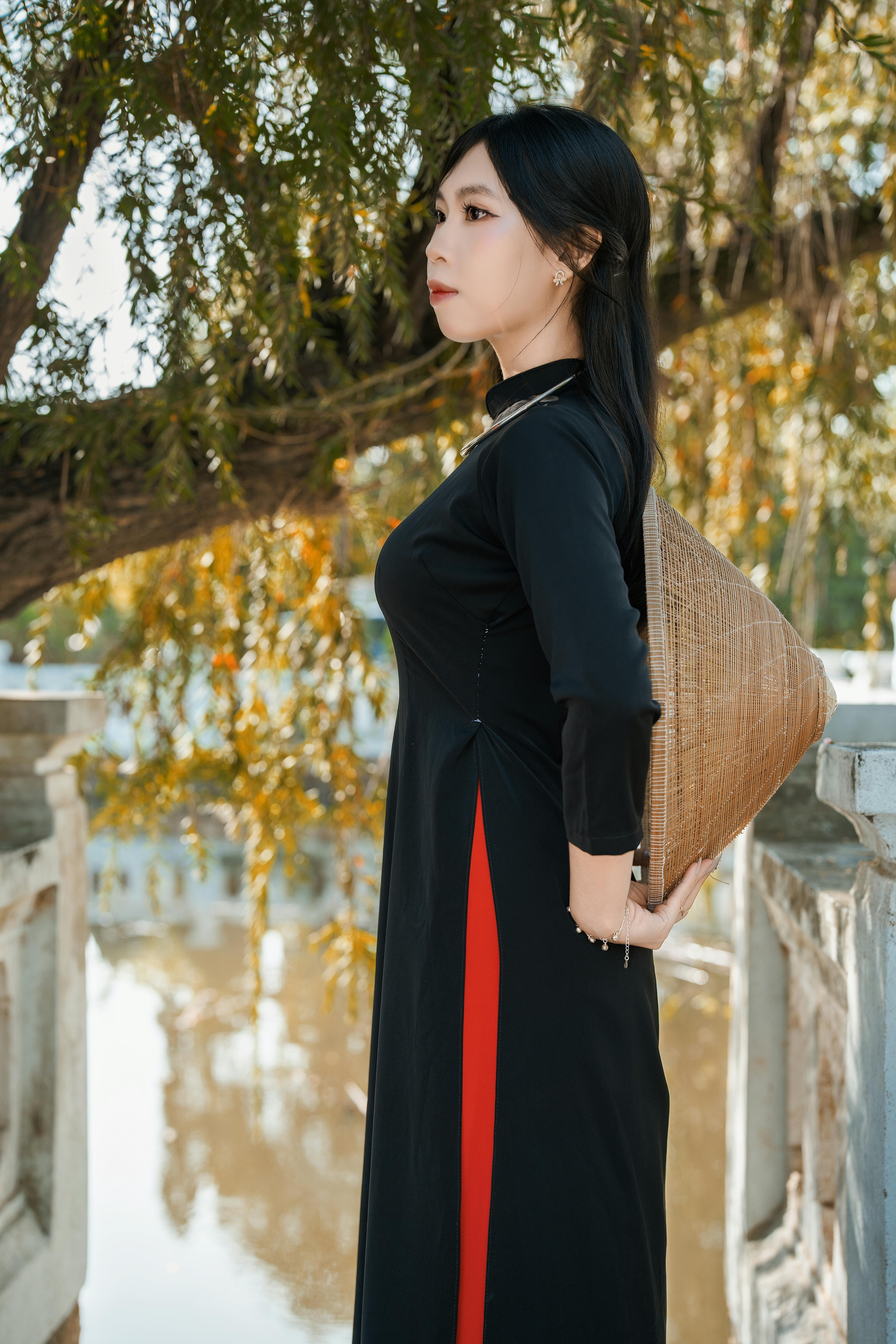 Side profile of a woman in a black ao dai, holding a conical hat, by a tranquil river.