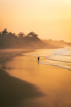 A solitary surfer walks along a golden beach at sunset, capturing serene coastal beauty.