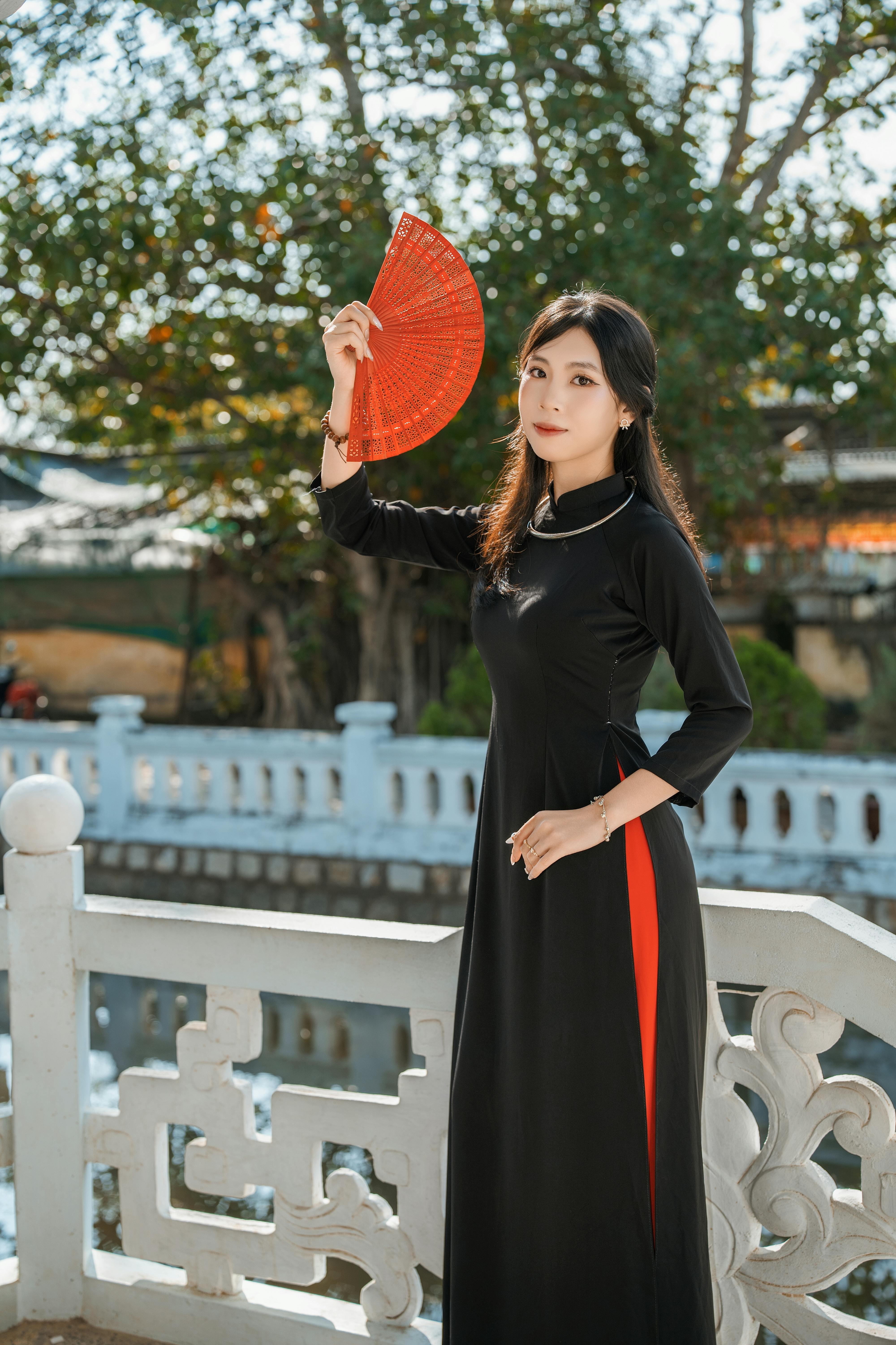 Asian woman in traditional attire gracefully holding a vibrant red fan outdoors, exuding elegance and cultural richness.