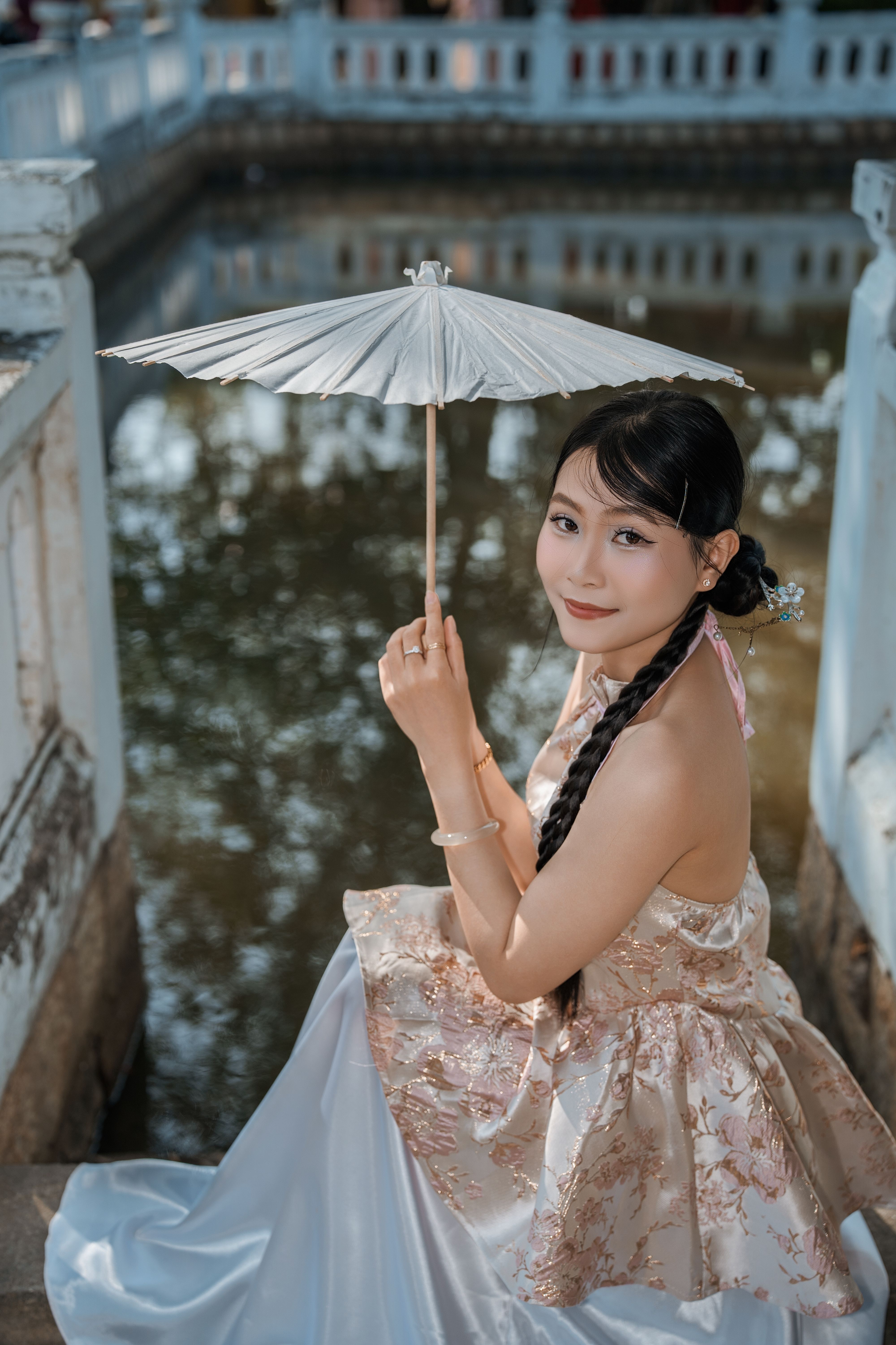 Woman with Asian parasol in elegant attire sitting by a serene pond, showcasing traditional style with a modern touch.