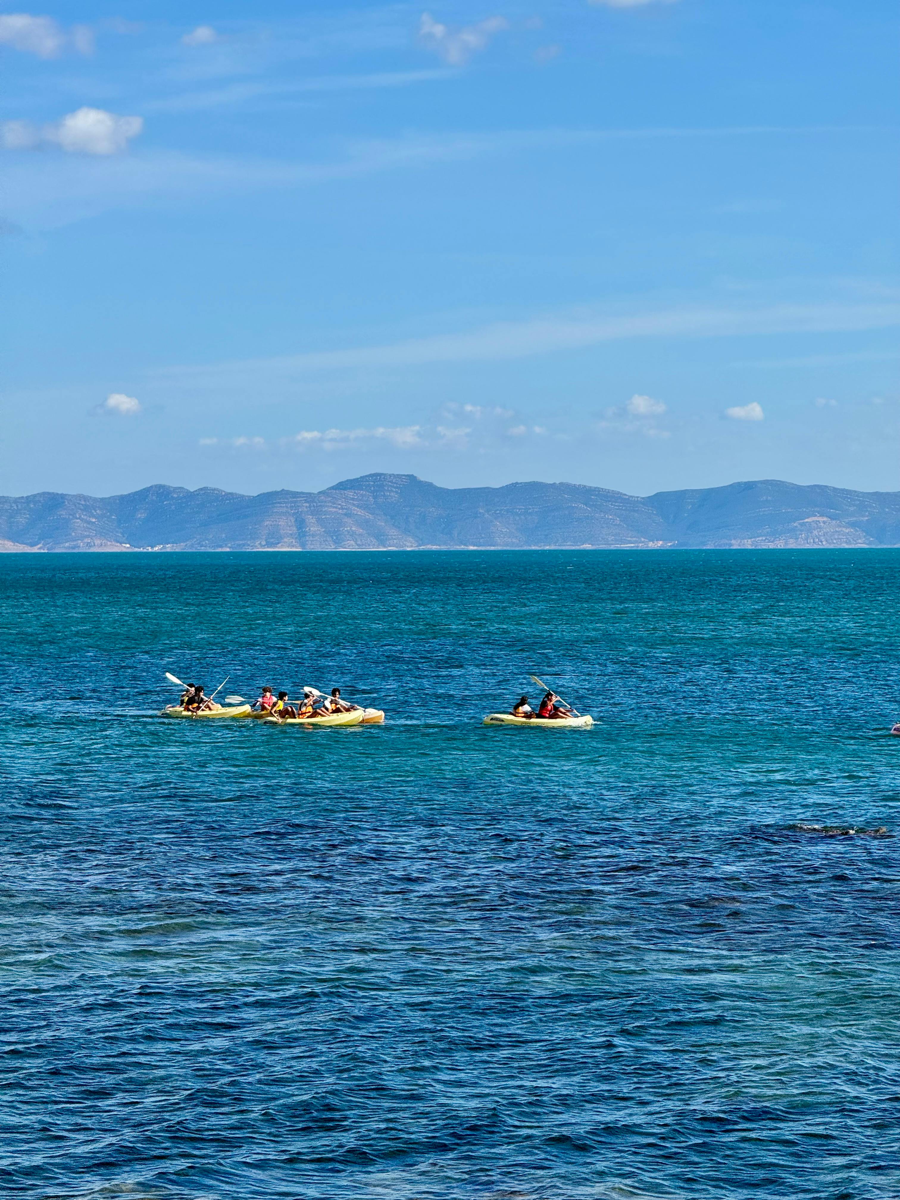 Kayakers paddle across the ocean with distant mountains under a clear sky.