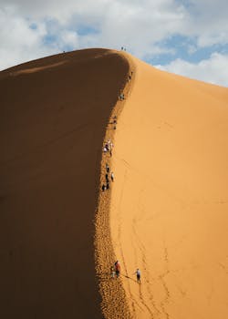Group of hikers ascending a large sand dune under blue sky, showcasing adventure and exploration.