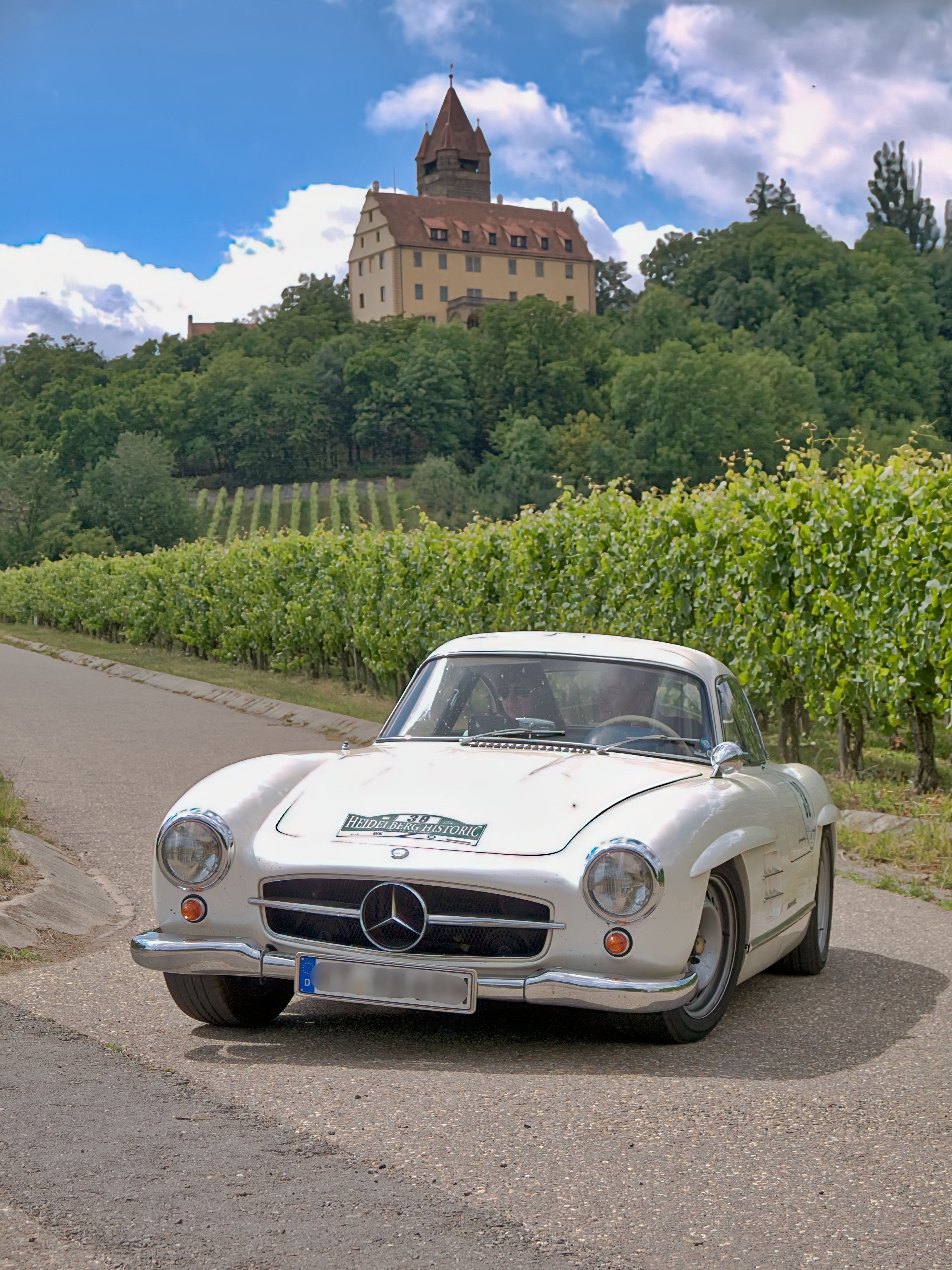 Classic Mercedes-Benz car parked along a scenic vineyard road near a historic castle.