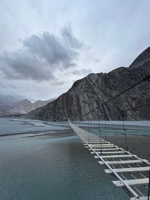 Free A suspension bridge spans a tranquil river against a dramatic mountainous backdrop under a cloudy sky. Stock Photo