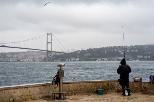 A fisherman stands by the Bosphorus, casting a line under a cloudy sky near the iconic bridge.