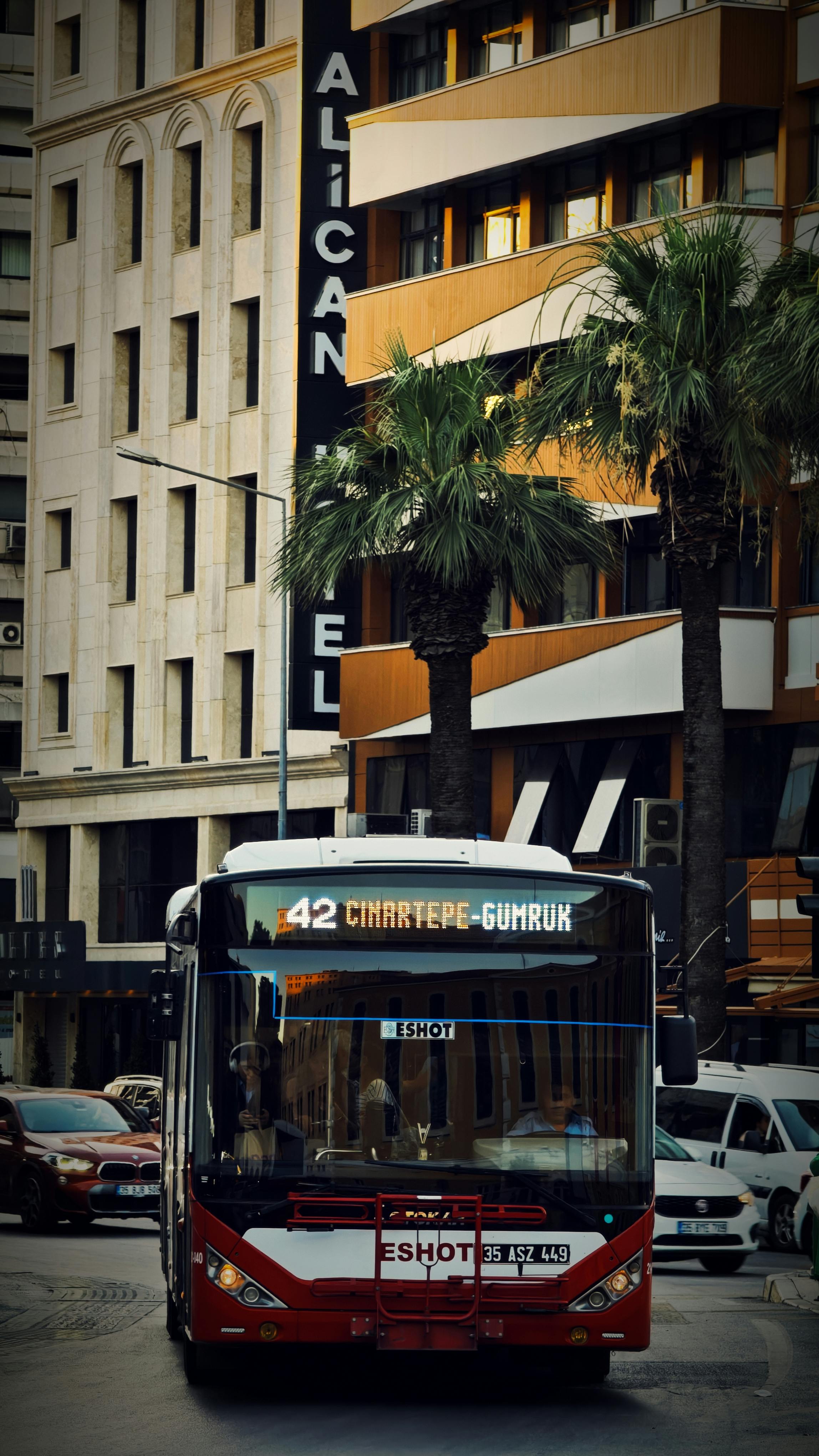 A city bus in İzmir, Türkiye, drives past palm trees and a modern building facade.