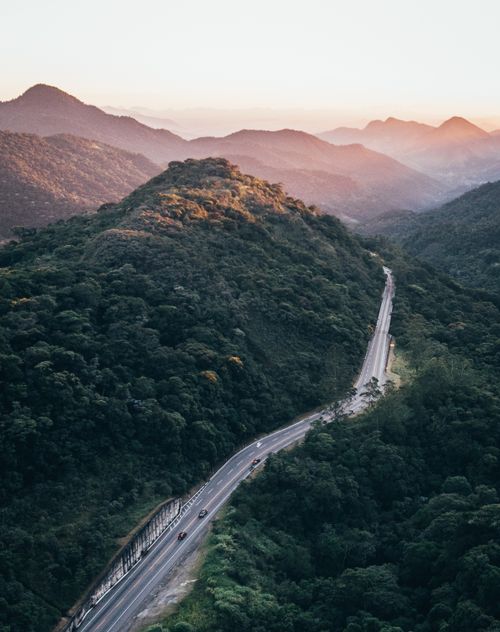 Free Drone captures a winding road through lush mountains at sunrise, offering stunning scenery. Stock Photo