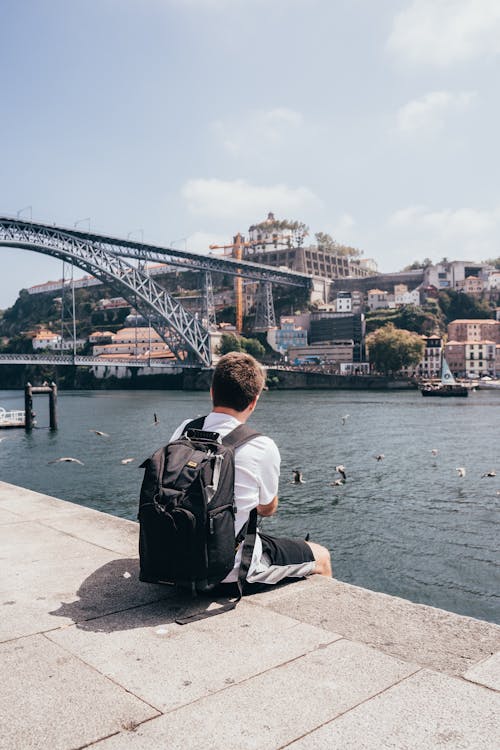 Back view of anonymous male tourist with backpack admiring cityscape on other side of river and openwork metal structure of bridge over river on sunny day