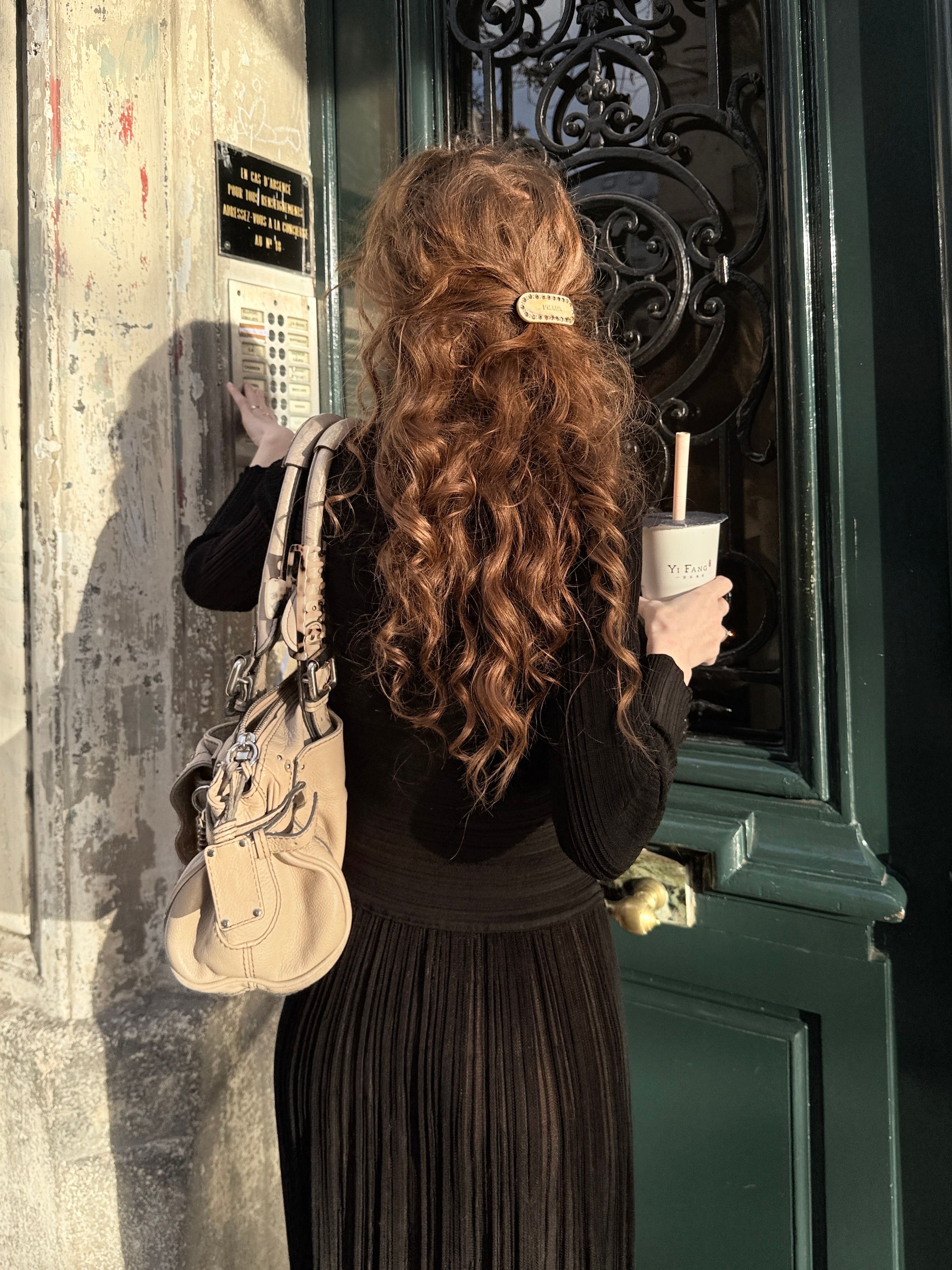 Woman with curly hair wearing a black dress standing at a doorway and holding a drink.