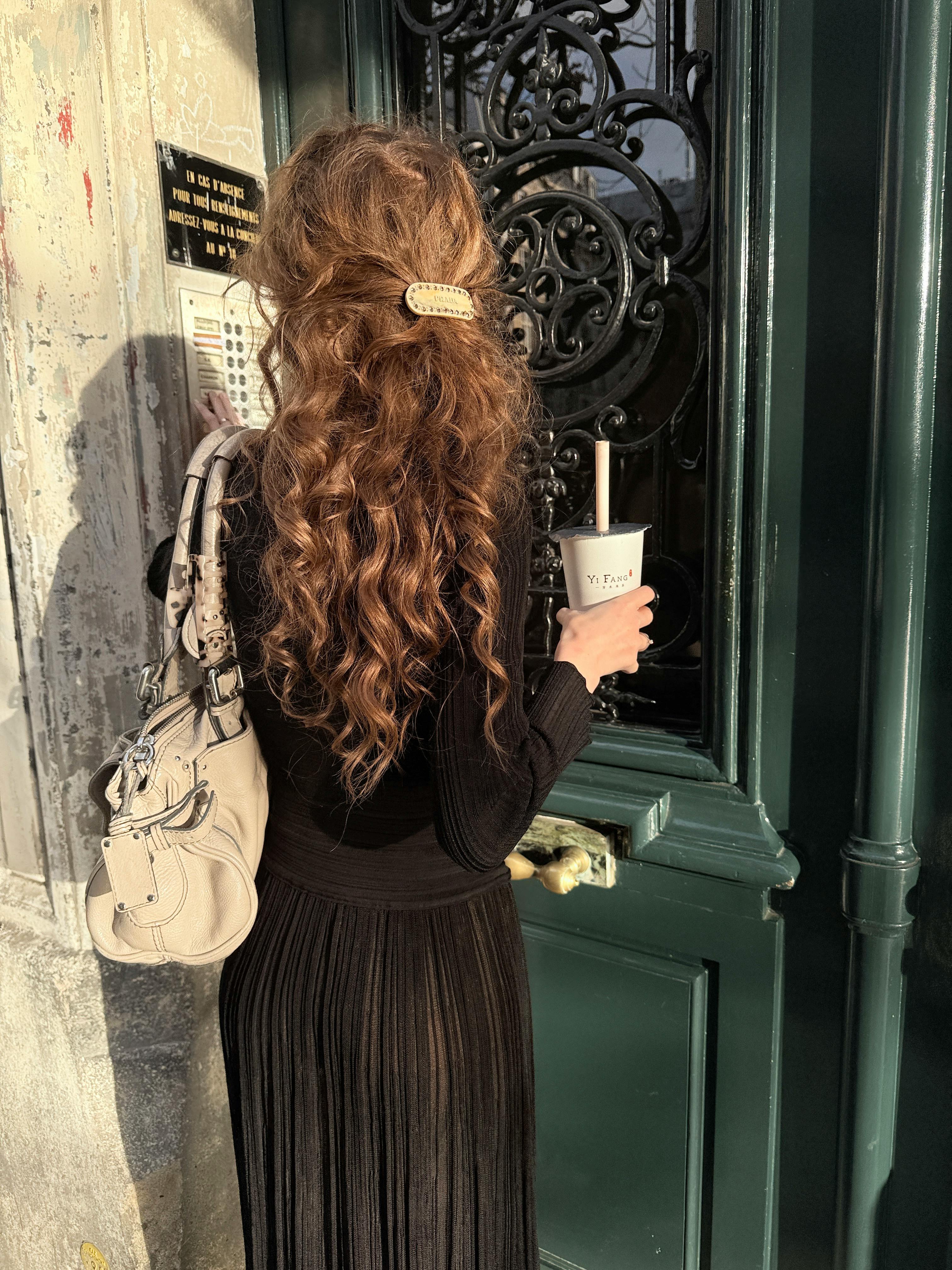 Woman with long hair entering a sunlit door holding a drink, exuding elegance.