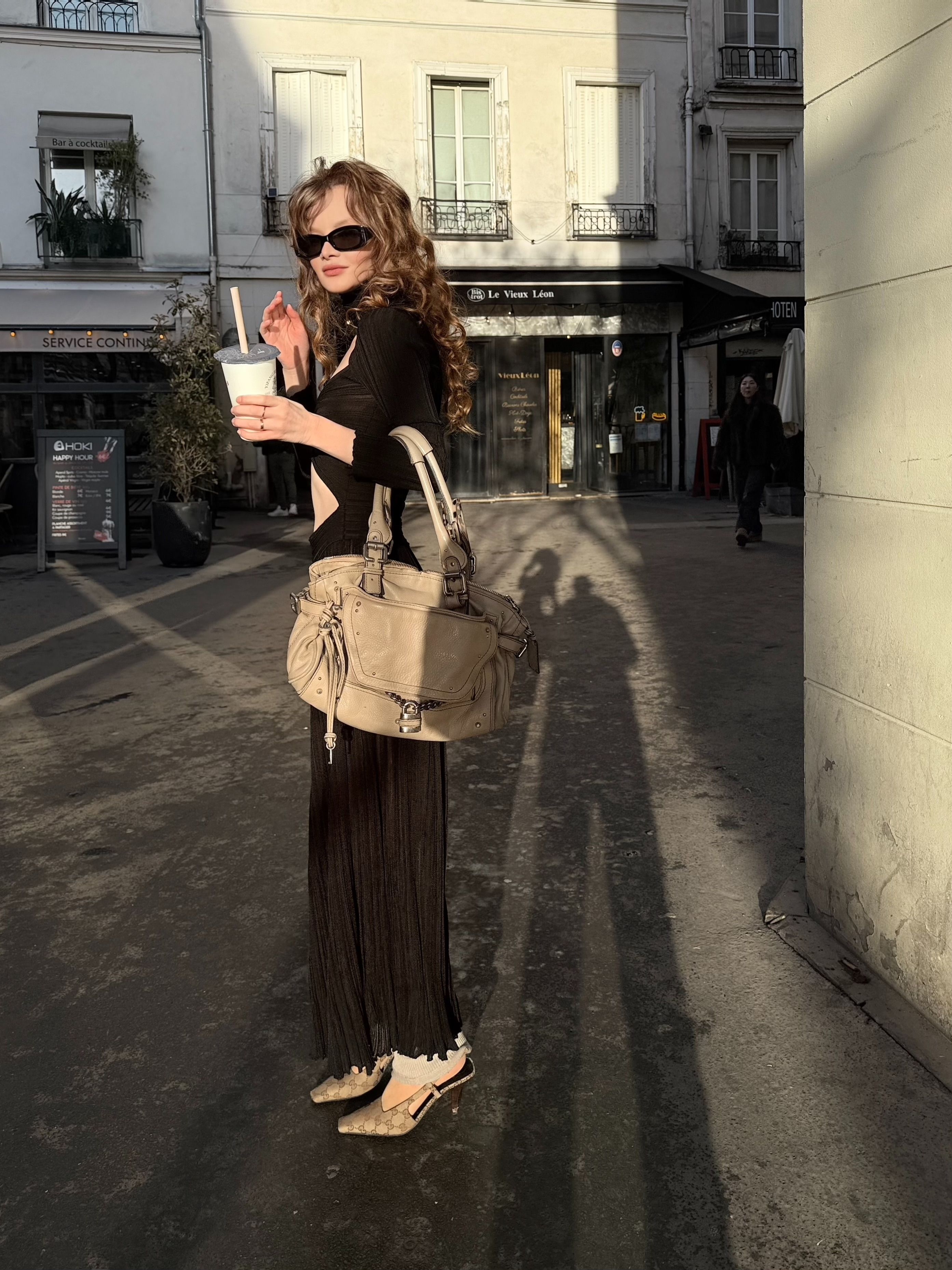 Woman in stylish black dress enjoys a drink in a sunlit city street.