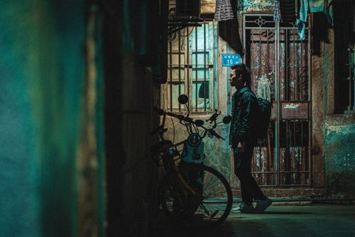 Free Side view of young Asian man in casual clothes with backpack walking near shabby house with bars on window and door in dark alley of aged city Stock Photo
