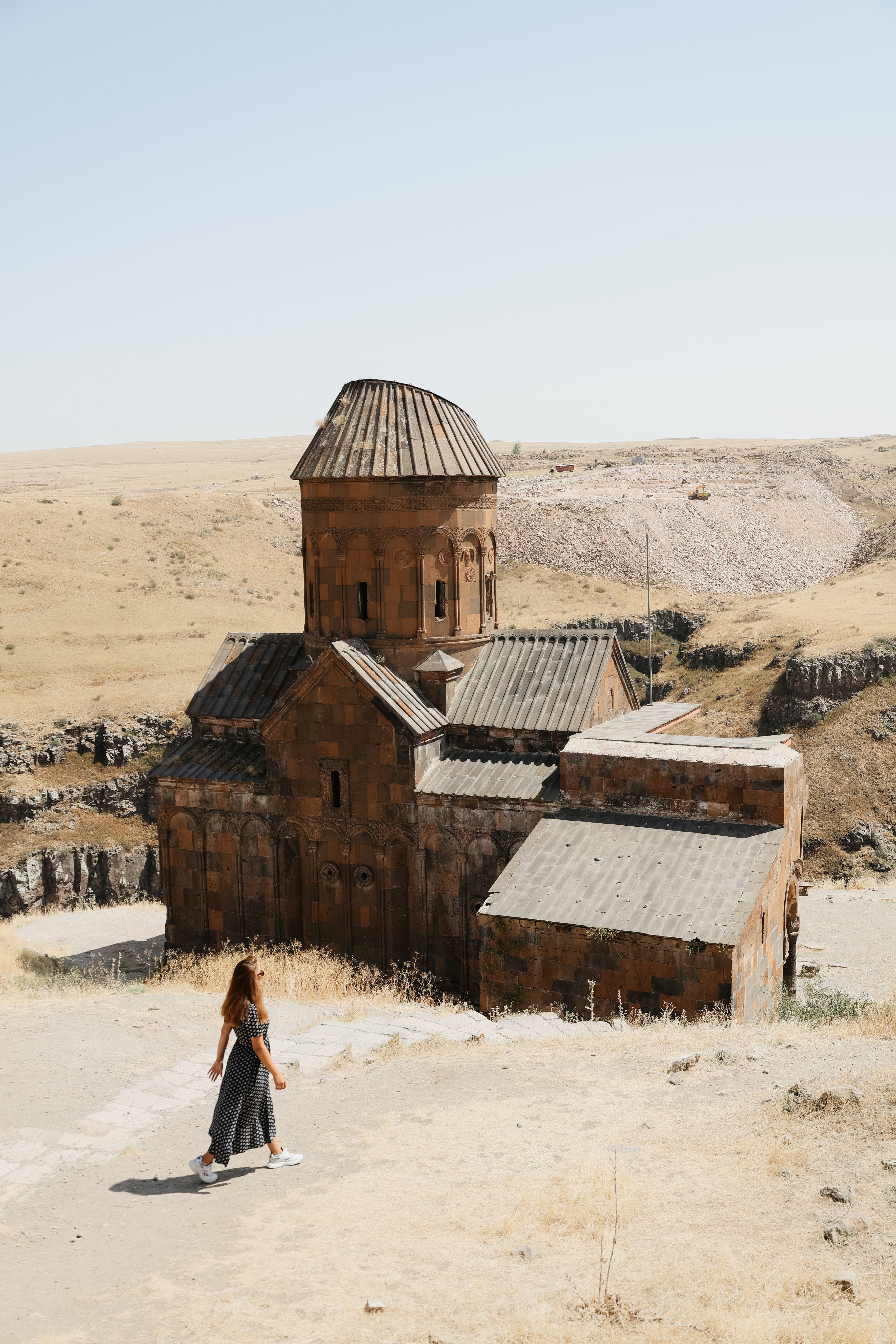 A woman walks near historic church ruins in a desert landscape under clear skies.