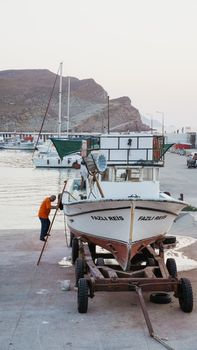 A fishing boat named Fazlı Reis docked in a tranquil harbor shortly after sunrise, with an individual making minor repairs.