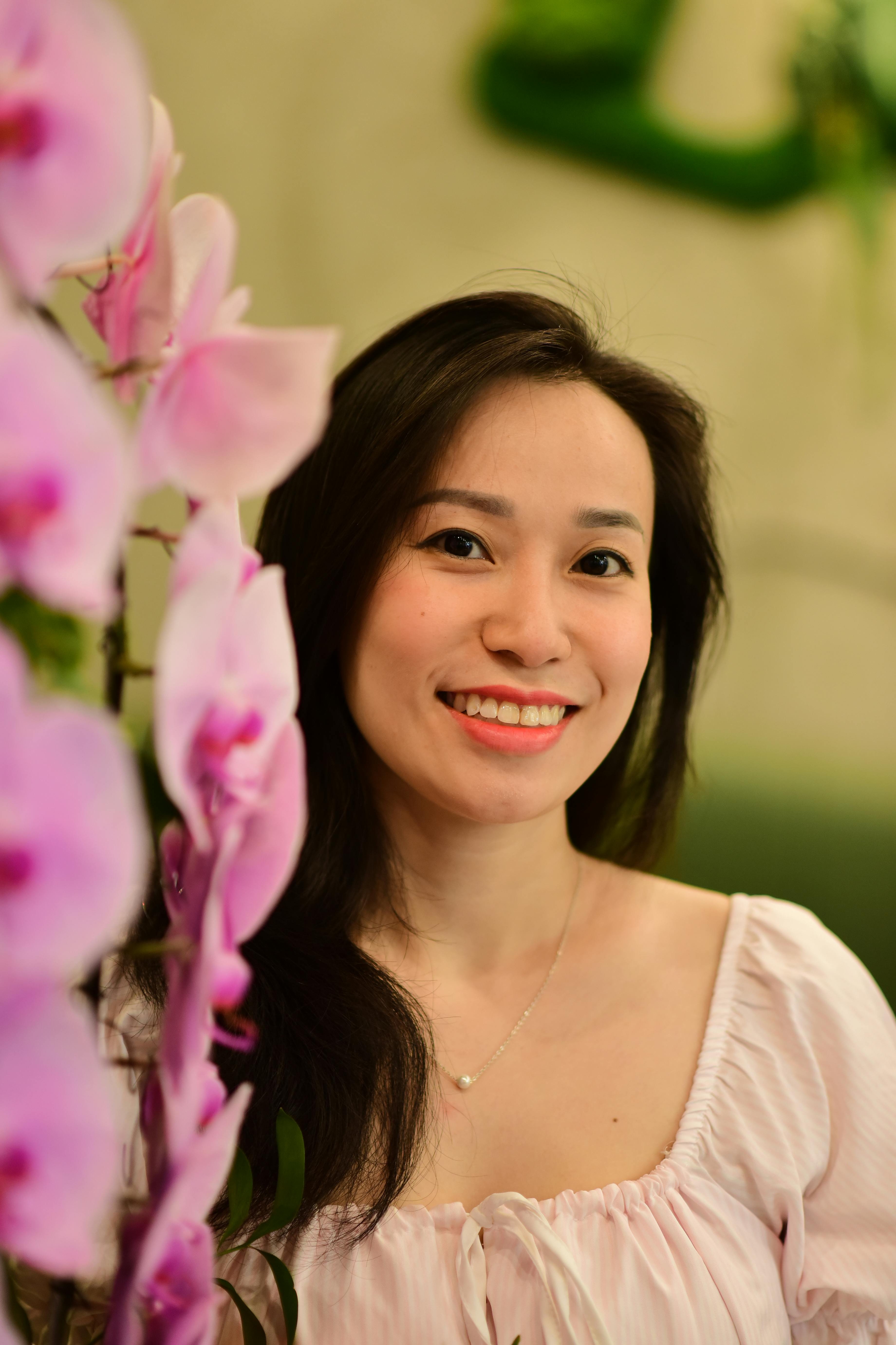 A cheerful woman posing beside vibrant pink orchids indoors.