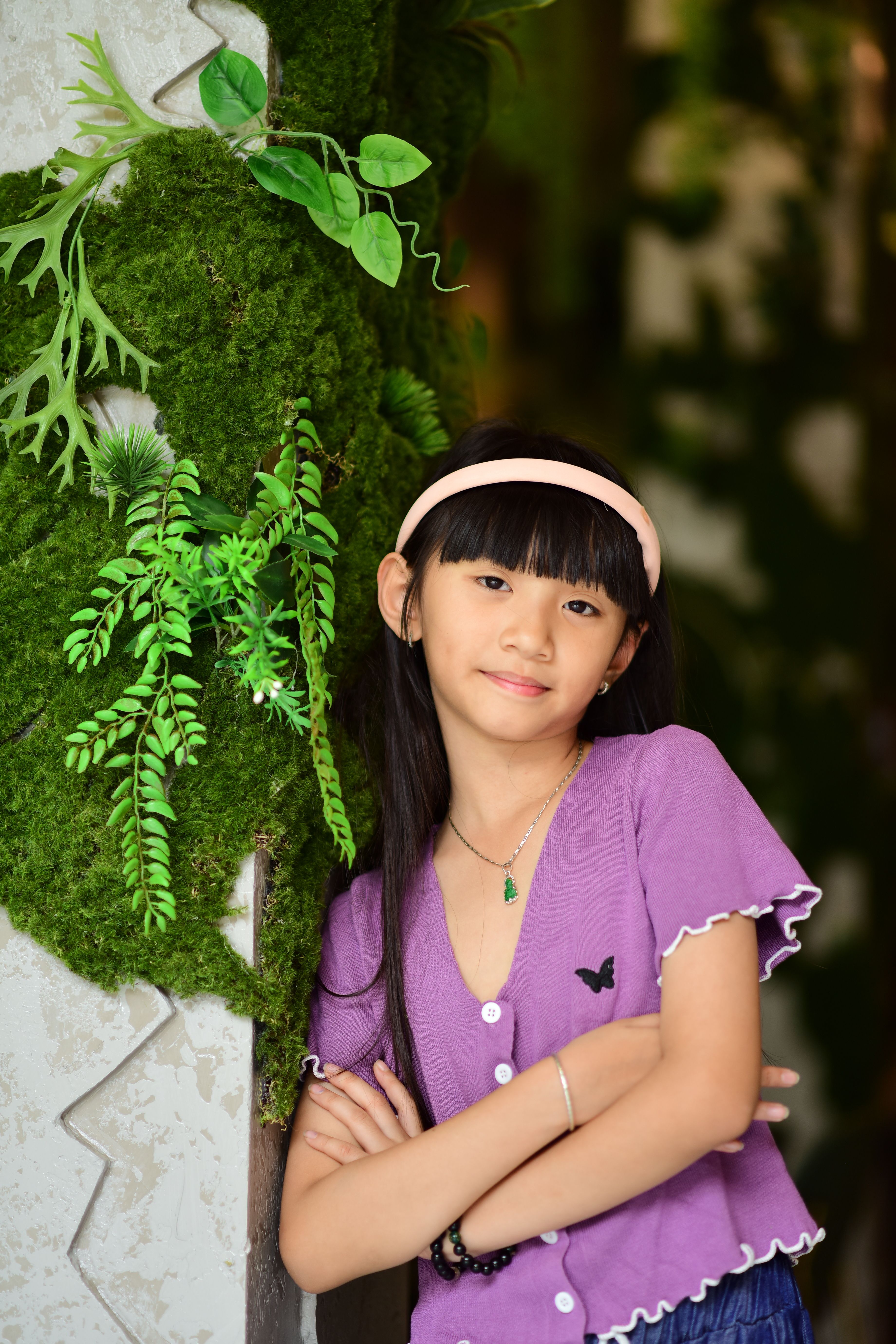 Young girl posing next to vibrant green foliage indoors, exuding confidence.