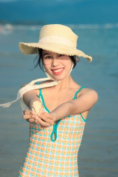 Smiling woman in a straw hat holds a seashell at the beach, exuding joyful summer vibes.