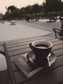 A serene outdoor café scene with a black coffee and book on a wooden table, perfect for a relaxing moment.