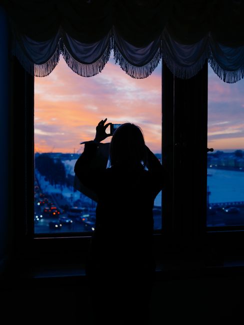 Silhouette of a woman photographing a vibrant sunset through a window with a scenic view.