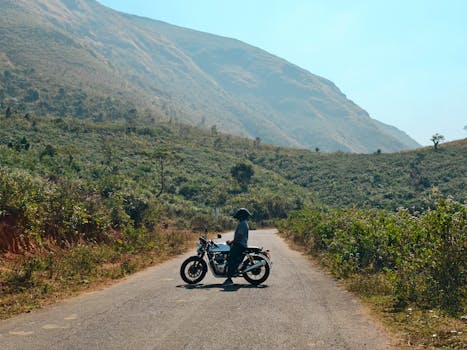 A motorcyclist rests on a scenic mountain road surrounded by lush greenery and hills.