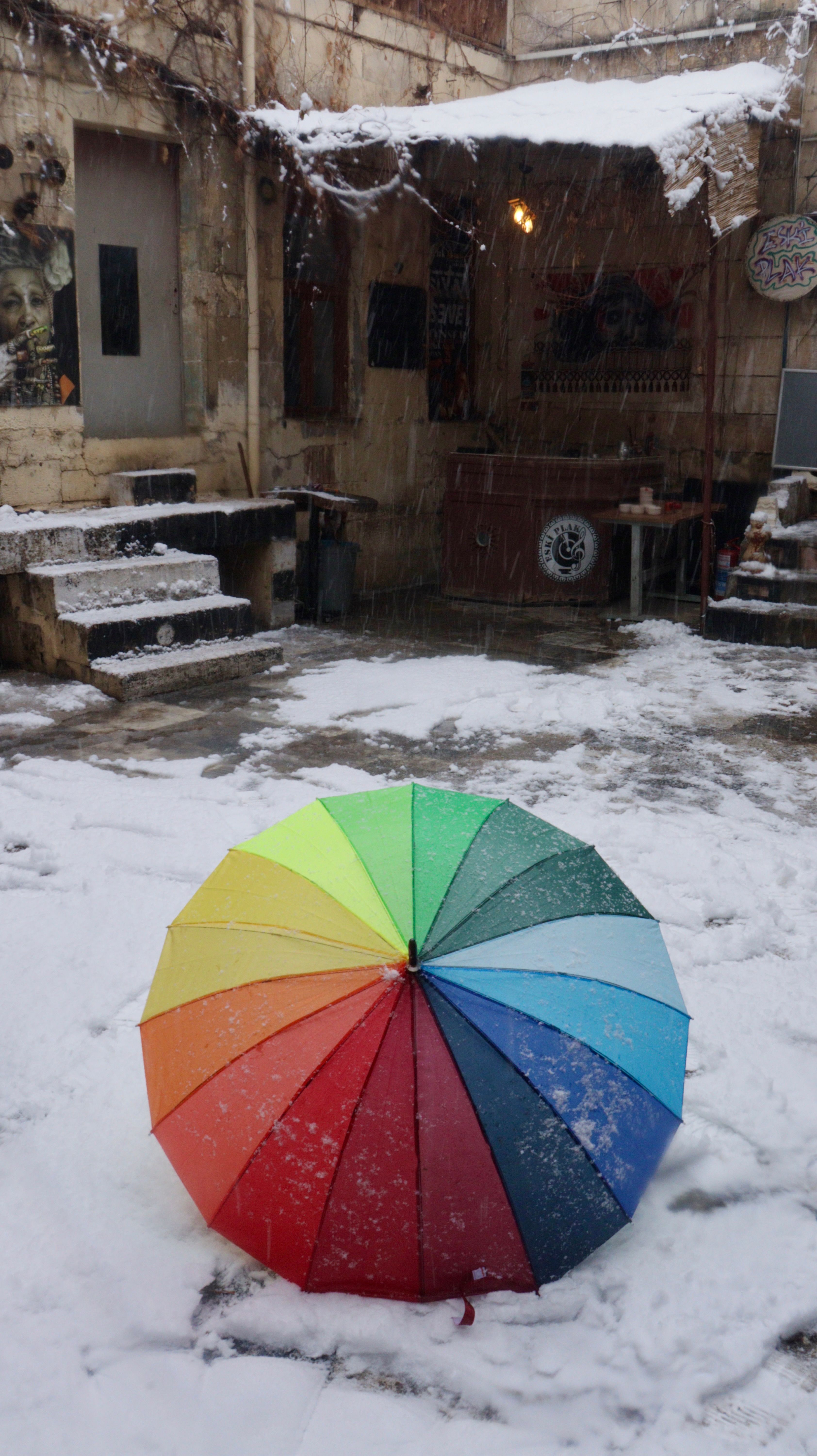 Vibrant rainbow umbrella in a snow-dusted rustic courtyard, creating a striking contrast.
