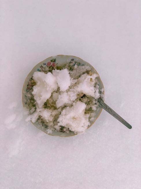A speckled white bowl of freshly made snow ice cream with a drizzle of vanilla, sitting on a snowbank outdoors with more falling snow in the background.