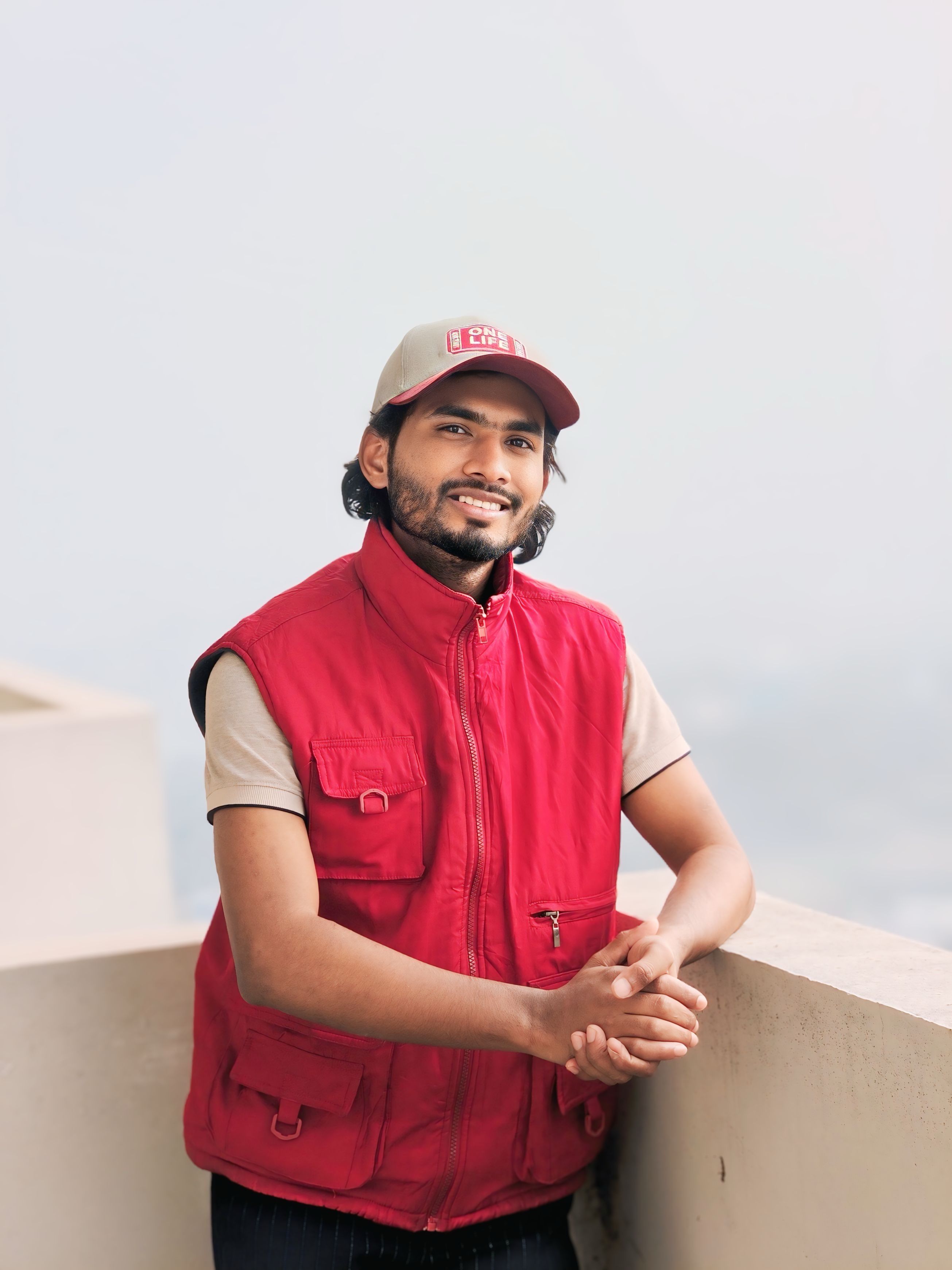 Confident man in a red vest and cap posing casually outdoors against a bright sky.