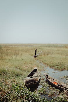 Scenic view of marshland with wooden boats and a solitary fisherman under clear sky.