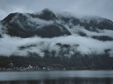 Misty mountains tower over a serene lakeside village, enveloped in clouds.