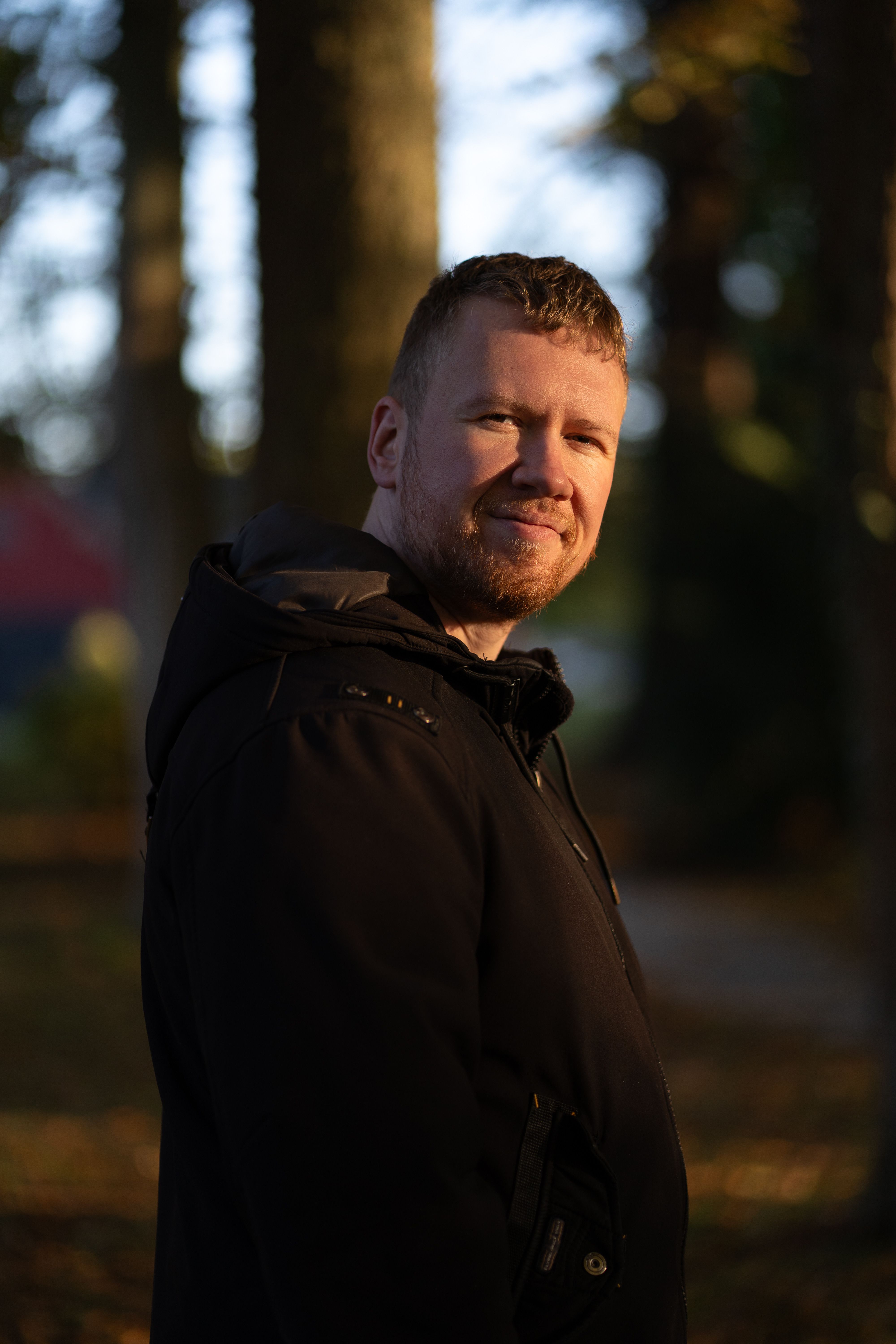 A man standing outdoors in a park during autumn with soft, warm sunlight.