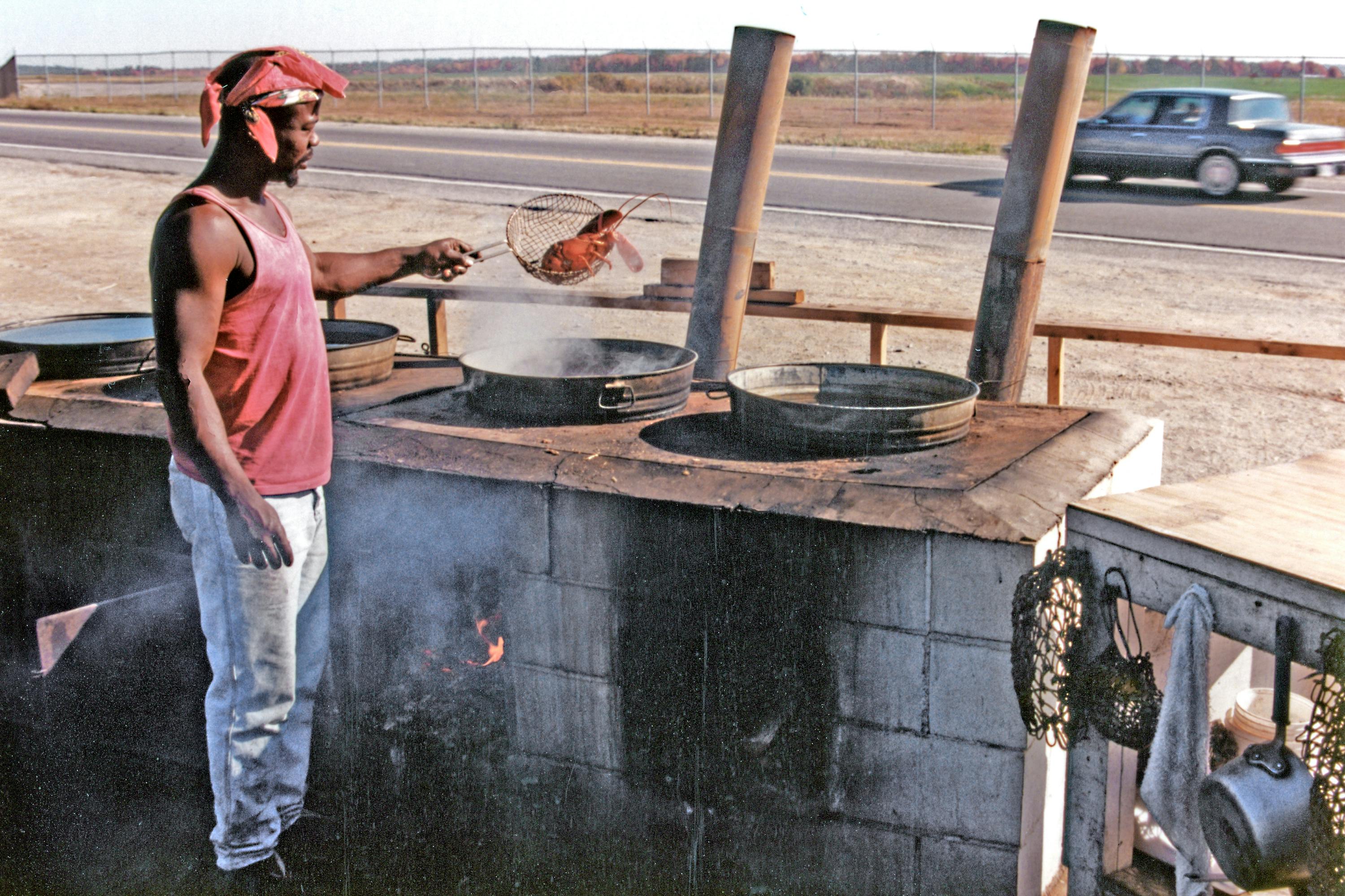 A man cooking Maine lobster outdoors by the roadside using traditional methods.