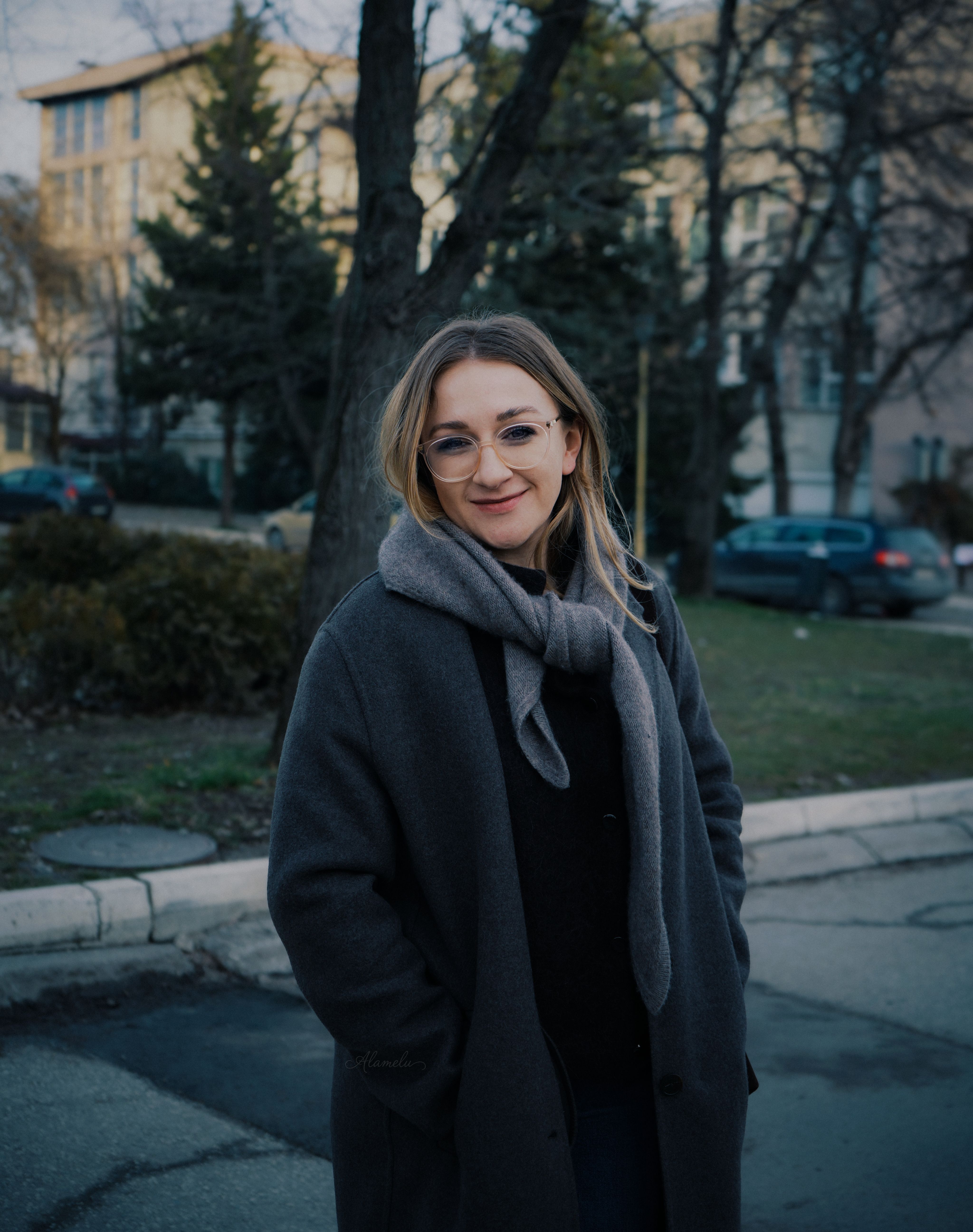 Smiling woman in a winter coat and glasses standing outdoors during the day.