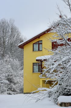 A bright yellow house stands amid a peaceful snow-covered landscape, evoking a winter wonderland.