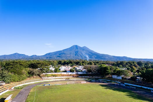 Aerial view of a sports stadium in Zacatecoluca, El Salvador, with a volcano in the background under a clear blue sky.