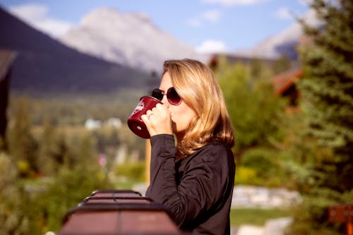 Shallow Focus Photo of Woman in Black Long-sleeved Shirt Drinking on Maroon Ceramic Mug