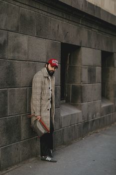 A fashionable man wearing a plaid coat and red cap stands against a textured urban wall.