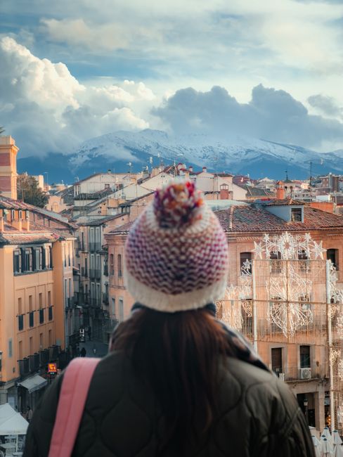 A woman gazes over a picturesque European town with snowy mountains in the background, under a dramatic sky.