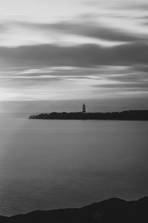 Free A tranquil black and white coastal scene featuring a distant lighthouse under dynamic skies. Stock Photo