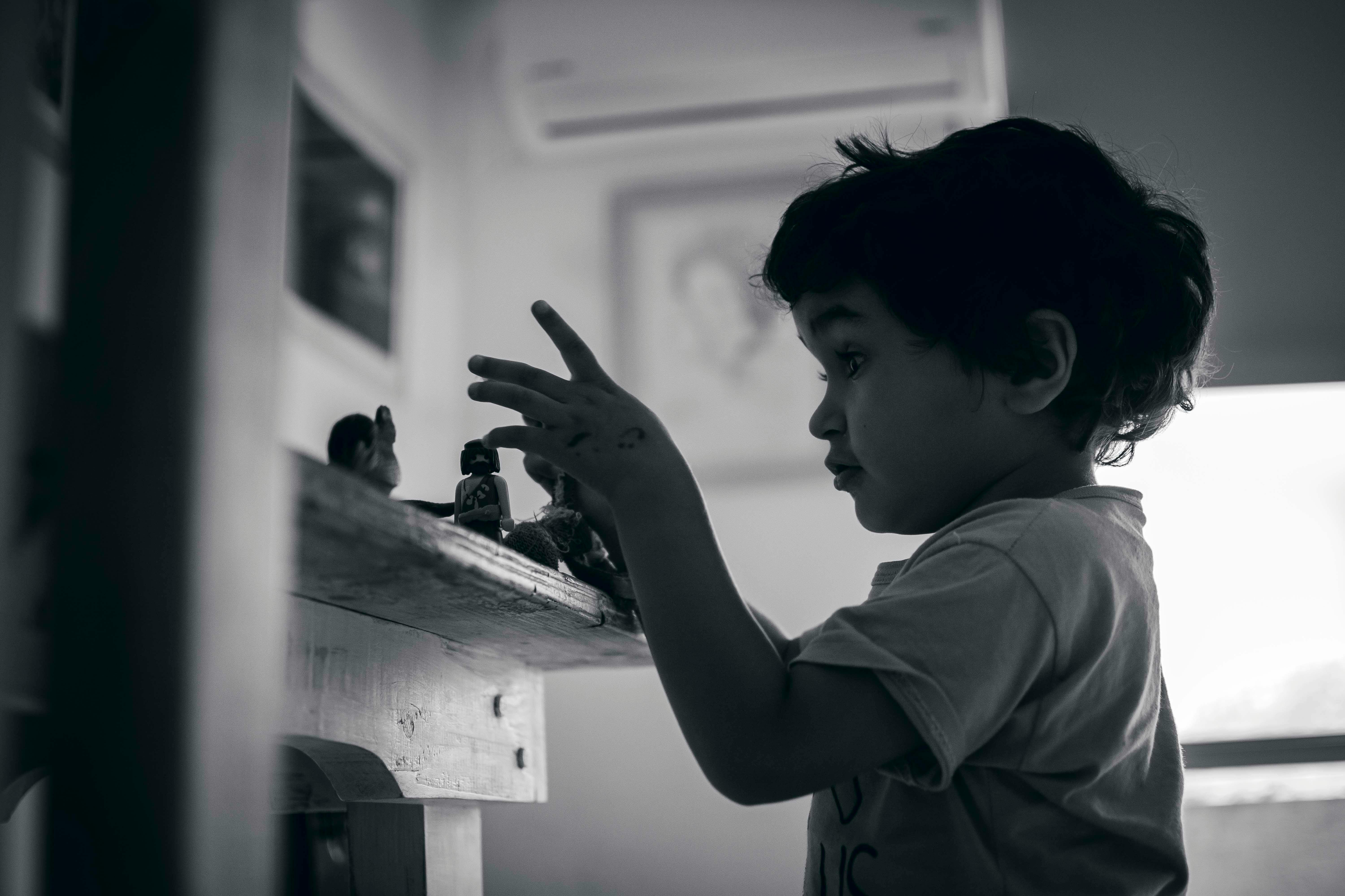A black and white image of a child engaging with toys on a shelf, captured indoors.