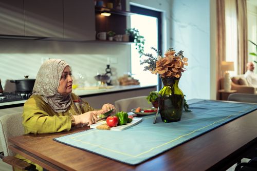 Free A Middle Eastern woman in a modern kitchen chopping vegetables. Bright ambiance, with flowers on the table. Stock Photo