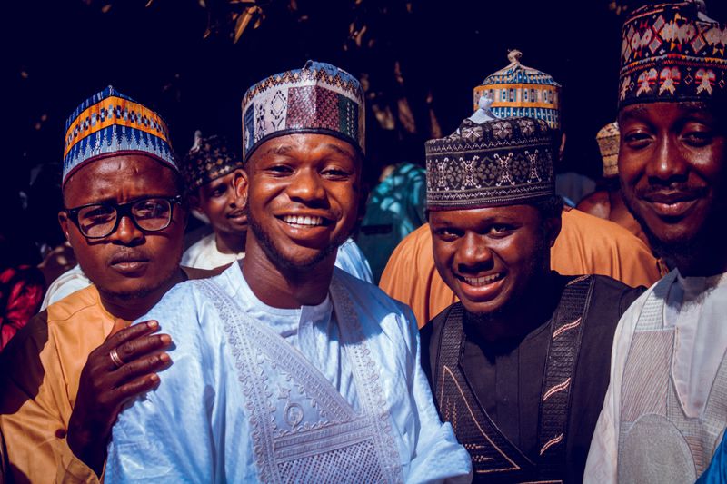 A cheerful group of Nigerian men wearing colorful traditional attire and caps, outdoors.