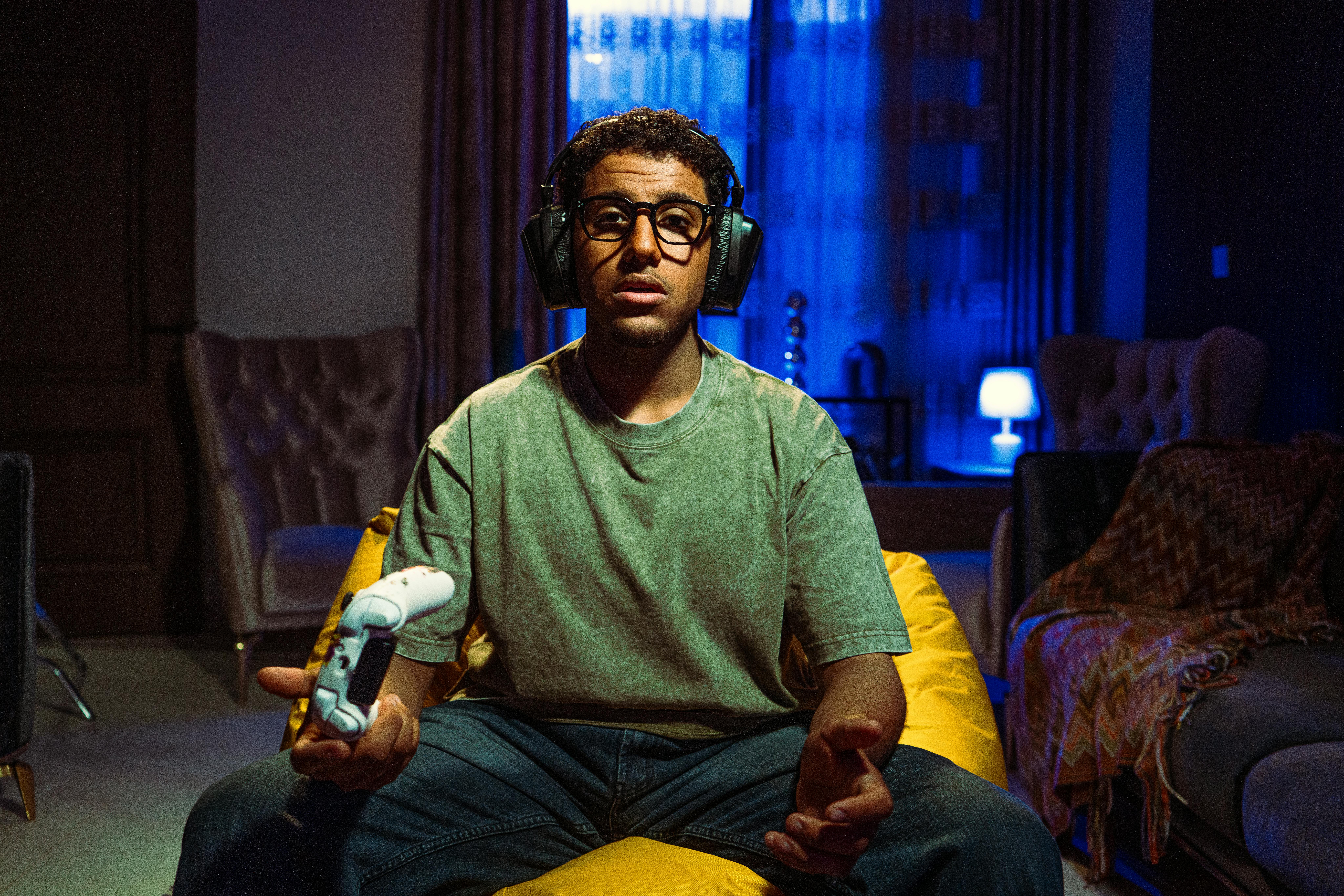 A man with headphones plays video games on a console in a cozy living room at night.