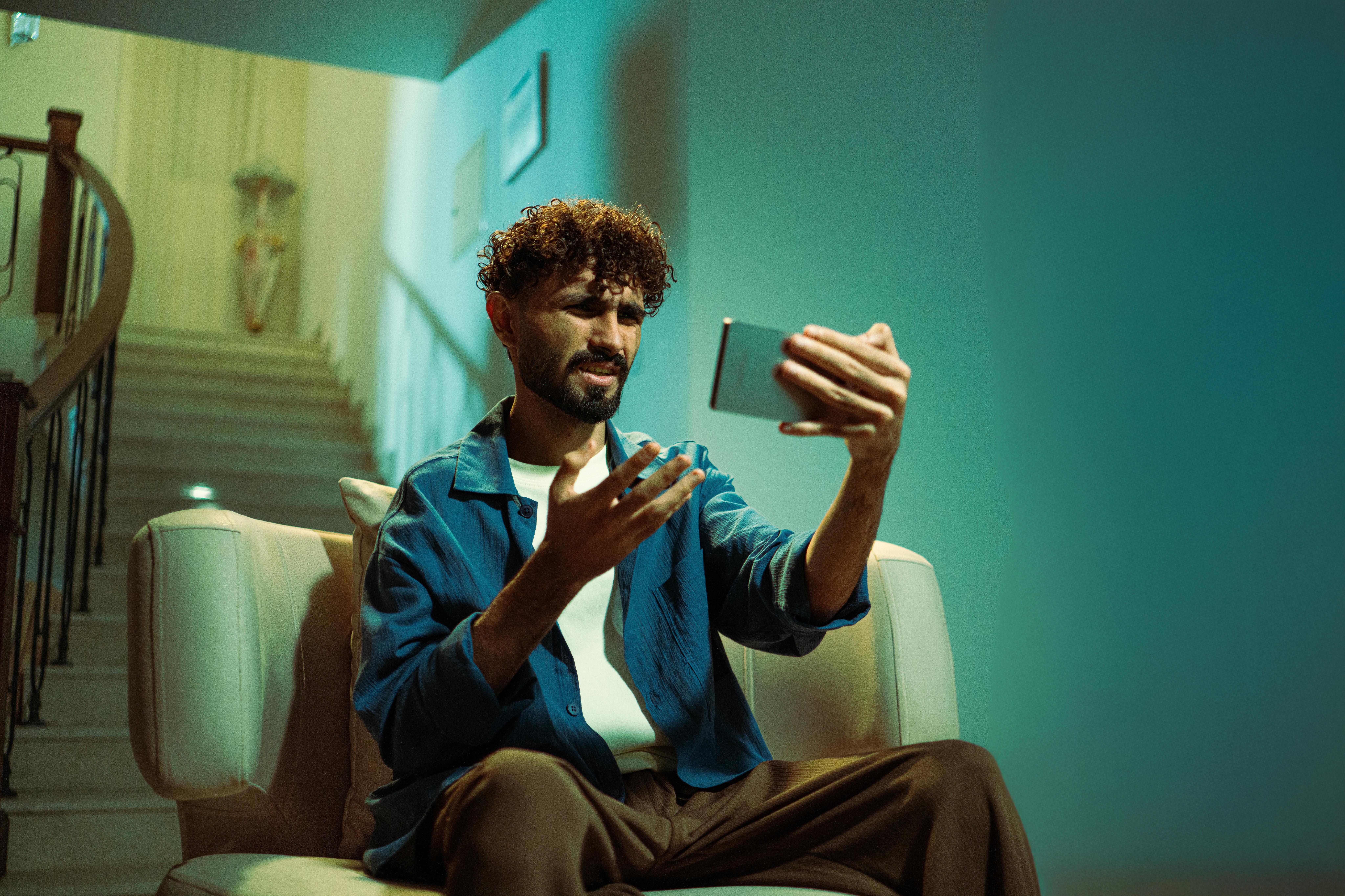 Miễn phí Một người đàn ông đang ngồi trên ghế sofa trong nhà, vẻ mặt bối rối trong khi sử dụng điện thoại thông minh. Ảnh lưu trữ