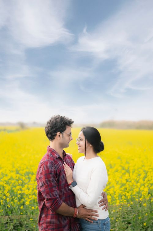 Free A smiling couple embraces in a vibrant field of yellow flowers under a clear blue sky. Stock Photo